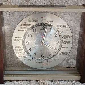 May include: A silver world clock with a black face and silver hands. The clock shows the time in different cities around the world. The clock is framed in a brown wood frame.