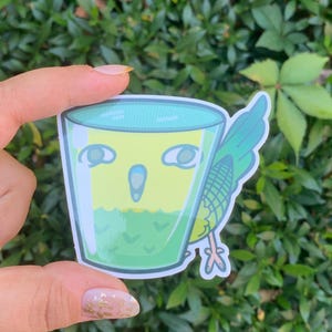 Könnte beinhalten: Ein grüner und gelber Aufkleber mit einem Cartoon-Vogel in einem Glas mit grüner Flüssigkeit. Der Vogel hat einen überraschten Ausdruck und schaut den Betrachter an.