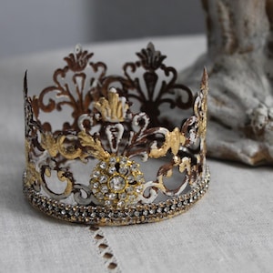 Pode incluir: Uma coroa decorativa com um design de filigrana. A coroa apresenta detalhes dourados, castanhos e brancos, com uma faixa de pequenos strass transparentes. A coroa é provavelmente usada para decoração ou como topo de bolo.