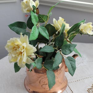 Puede incluir: Una tetera de cobre llena de rosas amarillas y hojas verdes. Las rosas están en plena floración y las hojas son de color verde oscuro. La tetera está sobre un mantel blanco con un patrón decorativo. El fondo es una pared gris claro y una ventana.