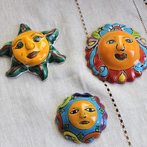 Könnte beinhalten: Drei farbenfrohe Wandbehänge aus Keramik mit Sonnenmotiven. Die Sonnen zeigen gelbe Gesichter mit ausdrucksstarken Merkmalen, umgeben von dekorativen Elementen in Grün-, Rot-, Blau- und Orangetönen. Die Sonnen haben unterschiedliche Formen und Größen.