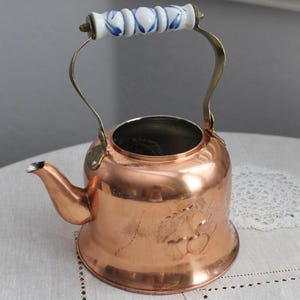 Pode incluir: Uma chaleira de cobre com bico e alça. A alça é feita de latão e tem uma pega de cerâmica branca e azul. A chaleira tem um design decorativo gravado na lateral.