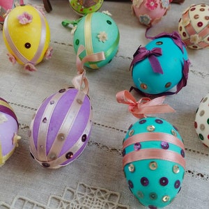 Op de afbeelding: Een verzameling decoratieve paaseieren in verschillende kleuren, versierd met linten, pailletten en bloemaccenten. Sommige eieren hebben geschilderde ontwerpen, waaronder een konijnenillustratie. Elk ei heeft een lintlus om op te hangen.