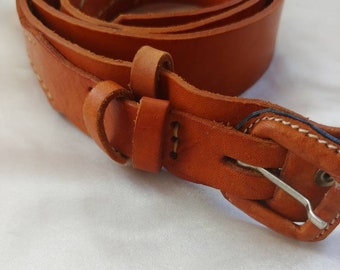 May include: A rolled-up, tan leather belt with a silver-colored buckle. The belt has white stitching along the edges and a loop to secure the belt end. The leather has a smooth texture and a warm, natural color.