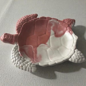 May include: A ceramic turtle-shaped dish with a pink and white color scheme. The dish features a textured shell and head, with the interior painted in a marbled effect. The dish is designed to resemble a turtle.