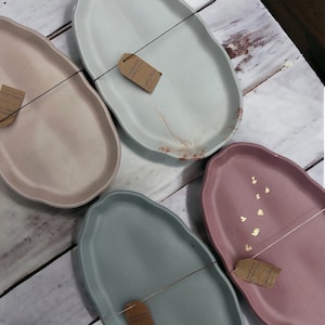 Peut inclure: Quatre plateaux en ciment faits à la main dans différentes couleurs. Les plateaux sont de forme ovale avec un bord légèrement irrégulier. Les plateaux sont étiquetés avec le texte "Handmade Cement Tray" et "Lumelite Ce".