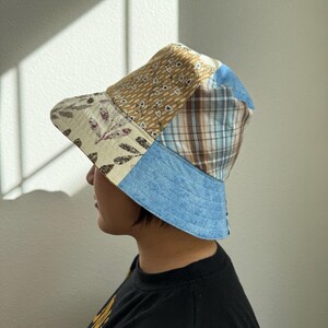 May include: A patchwork bucket hat featuring a variety of fabrics in beige, blue, and brown. The hat has a light blue brim and a mix of patterned and solid fabrics on the crown, creating a unique, handcrafted look. The hat is worn by a person.