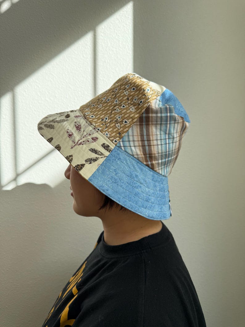May include: A patchwork bucket hat featuring a variety of fabrics in beige, blue, and brown. The hat has a light blue brim and a mix of patterned and solid fabrics on the crown, creating a unique, handcrafted look. The hat is worn by a person.