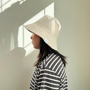 May include: A beige and white gingham bucket hat with a dark blue brim. The hat is worn with a black and white striped t-shirt. The hat has a small white tag on the side. The person is facing left.