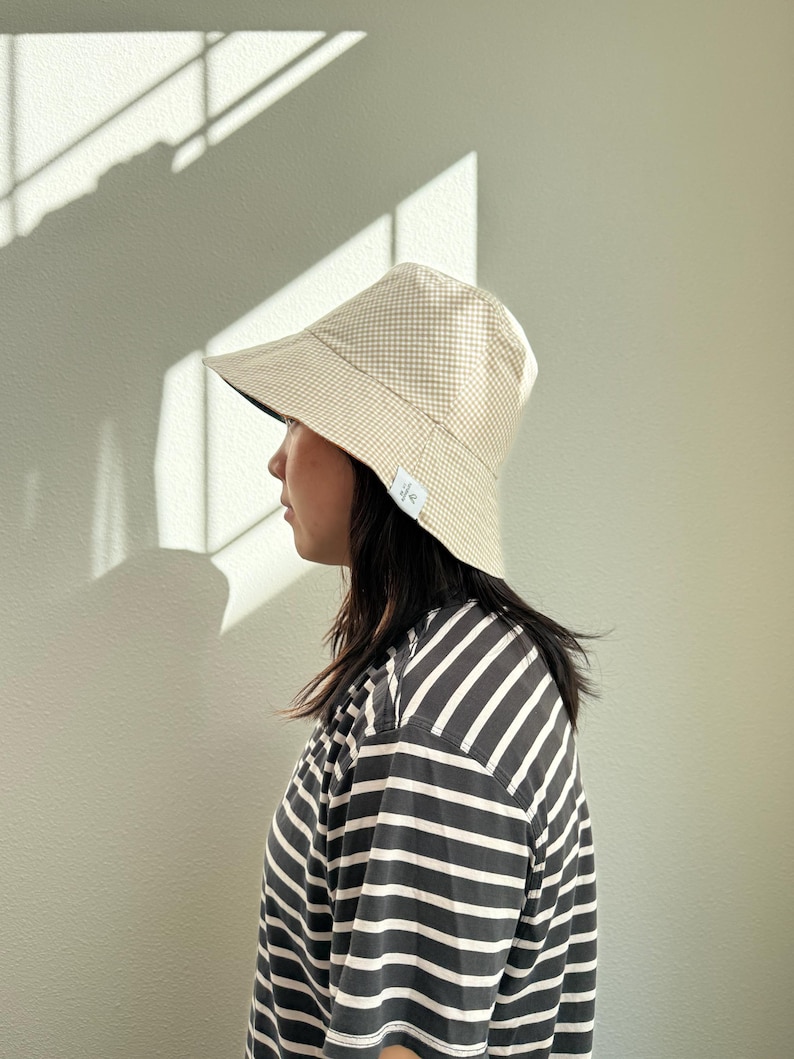 May include: A beige and white gingham bucket hat with a dark blue brim. The hat is worn with a black and white striped t-shirt. The hat has a small white tag on the side. The person is facing left.