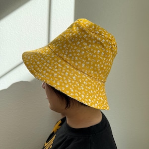 May include: A yellow bucket hat with a white floral pattern. The hat is made of fabric and has a wide brim. The hat is worn by a person wearing a black shirt.