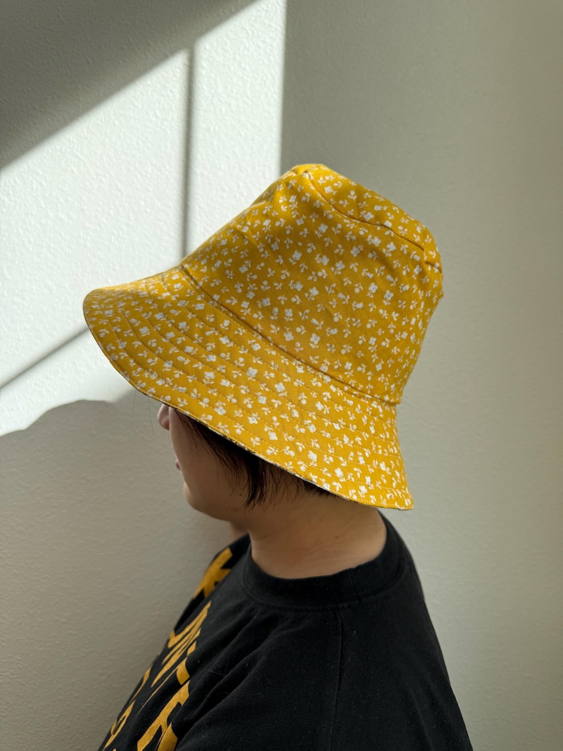 May include: A yellow bucket hat with a white floral pattern. The hat is made of fabric and has a wide brim. The hat is worn by a person wearing a black shirt.