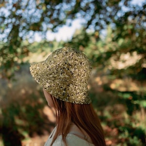 Op de afbeelding: Een persoon draagt ​​een groene en bruine gebloemde bucket hat. De hoed heeft een brede rand en is gemaakt van een zachte, lichtgewicht stof.