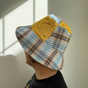 Può includere: Un cappello a secchiello patchwork con un mix di motivi e colori. Il cappello combina una stampa floreale gialla con un design a quadri blu, marrone e bianco. Un accessorio elegante per la protezione solare.