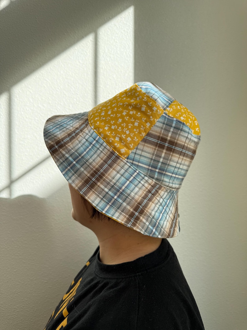 May include: A patchwork bucket hat featuring a mix of patterns and colors. The hat combines a yellow floral print with a blue, brown, and white plaid design. The hat is a stylish accessory for sun protection.