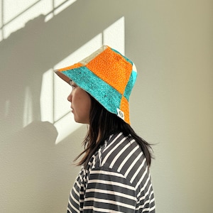 May include: A colorful patchwork bucket hat with orange, teal, and cream panels. The hat is worn by a person wearing a black and white striped shirt. The hat has a small logo on the side.