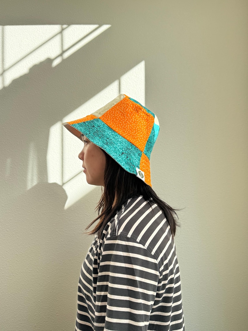 May include: A colorful patchwork bucket hat with orange, teal, and cream panels. The hat is worn by a person wearing a black and white striped shirt. The hat has a small logo on the side.