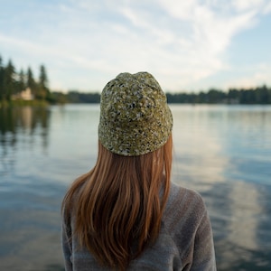 May include: A person wearing a green and white patterned bucket hat, looking out at a lake. The person has long brown hair and is wearing a grey sweater.