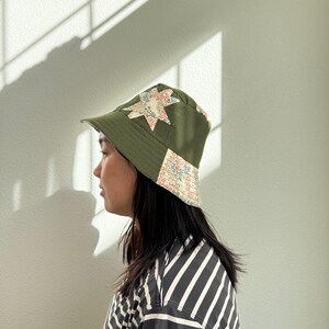 May include: Olive green bucket hat with star-shaped patchwork in floral and patterned fabric. The hat has a brim and is worn by a person. The hat is a stylish accessory.