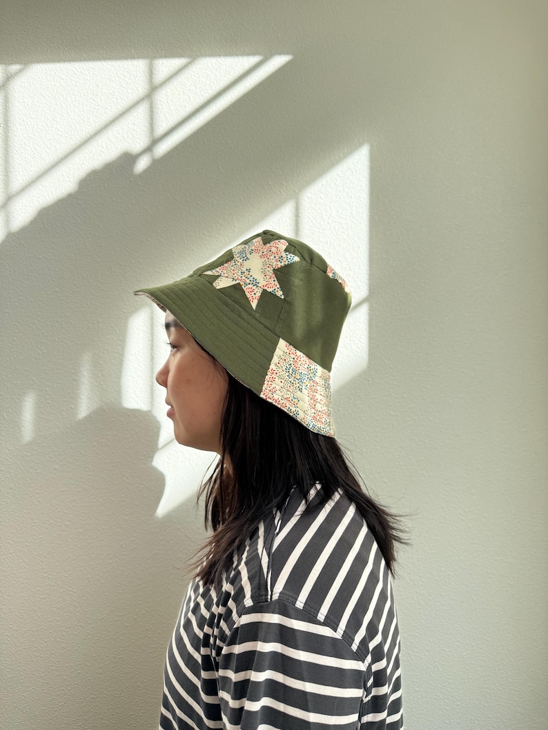 May include: Olive green bucket hat with star-shaped patchwork in floral and patterned fabric. The hat has a brim and is worn by a person. The hat is a stylish accessory.