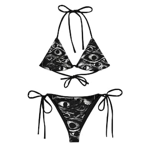 Könnte beinhalten: Ein zweiteiliger Bikini in Schwarz-Weiß mit einem sich wiederholenden Augenmuster. Das Oberteil ist im Neckholder-Stil mit Bindebändern, die Hose im klassischen Design mit Bindebändern an den Seiten. Der Badeanzug ist aus glattem Stoff.