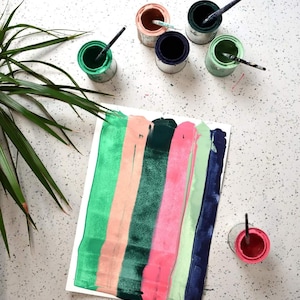 May include: A white sheet of paper with vertical stripes of green, peach, dark green, pink, light green, and dark blue paint.  There are six small paint cans with brushes on a white speckled surface.