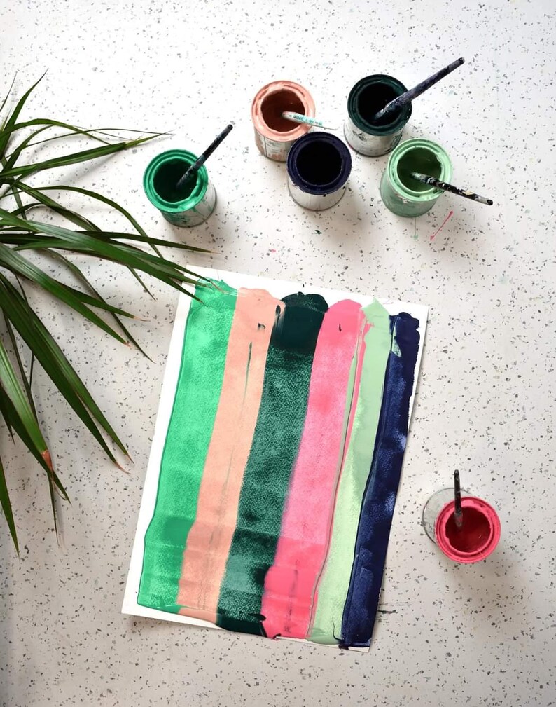 May include: A white sheet of paper with vertical stripes of green, peach, dark green, pink, light green, and dark blue paint.  There are six small paint cans with brushes on a white speckled surface.