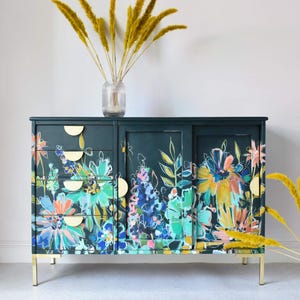 May include: A dark teal sideboard with a vibrant floral design in shades of blue, green, orange, and yellow. The cabinet has gold-colored hardware and legs. A clear glass vase with dried yellow grasses sits on top.
