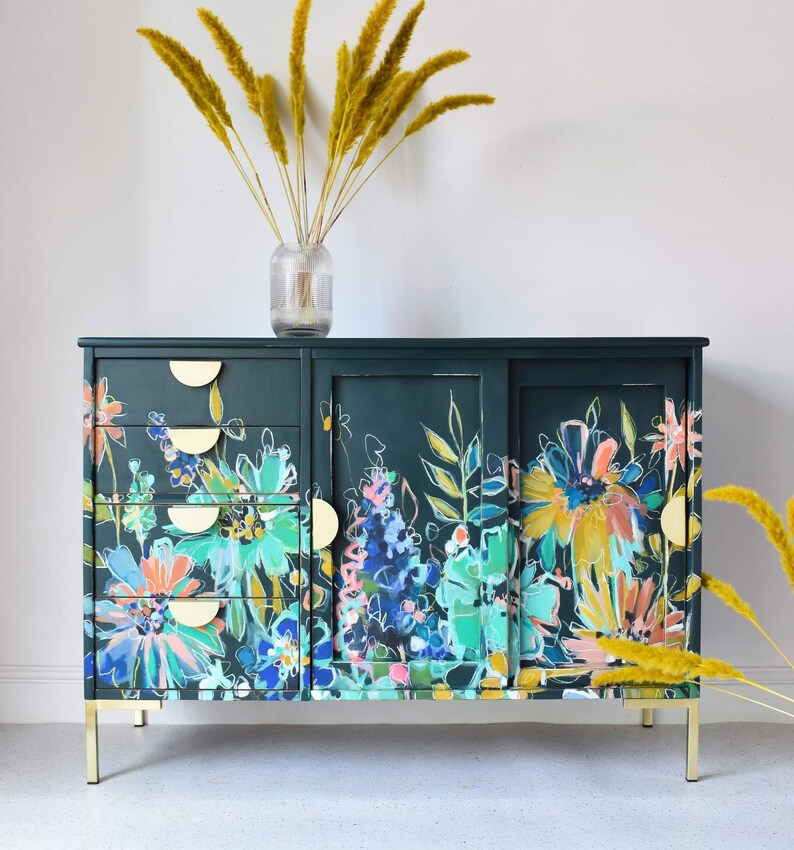 May include: A dark teal sideboard with a vibrant floral design in shades of blue, green, orange, and yellow. The cabinet has gold-colored hardware and legs. A clear glass vase with dried yellow grasses sits on top.