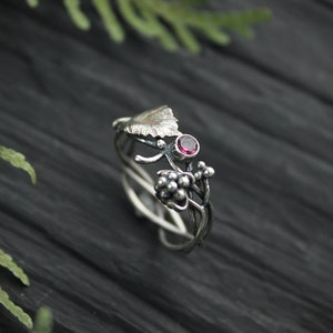 May include: A silver ring with a delicate design featuring a leaf, berries, and a pink gemstone. The ring is set against a dark wooden background.