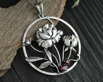 Può includere: Una collana con un ciondolo in argento con un designo circolare con due peonie stilizzate e foglie. Il ciondolo è accentato da due pietre preziose rosse.