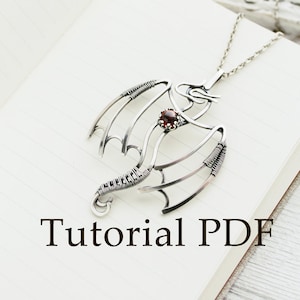 May include: A silver wire wrapped dragon pendant with a red gemstone, hanging on a silver chain. The pendant is on a white notebook with the text "Tutorial PDF".