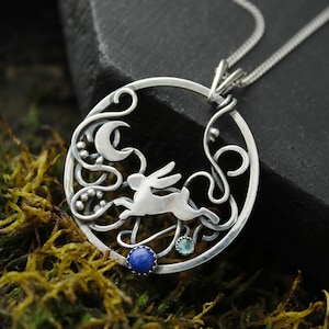 May include: A silver pendant necklace featuring a detailed design of a rabbit jumping through a moon and vines. The pendant has blue and green gemstone accents.