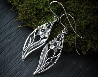 May include: A pair of silver-toned earrings featuring a floral design. Each earring has a stylized flower with a dark center, set within a leaf-shaped frame with delicate vine-like details. The earrings are displayed against a backdrop of green moss and a dark surface.
