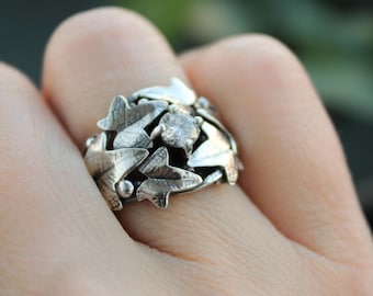 May include: A silver ring featuring a nature-inspired design. The ring is adorned with detailed leaf motifs and a central, clear gemstone. The leaves are intricately carved, creating a textured surface. The ring is shown on a finger, highlighting its size and design.