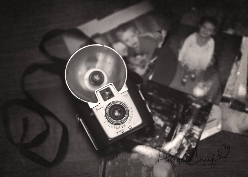 May include: A vintage black and silver camera with a flash attachment. The camera is sitting on a table with a black and white photo in the background.