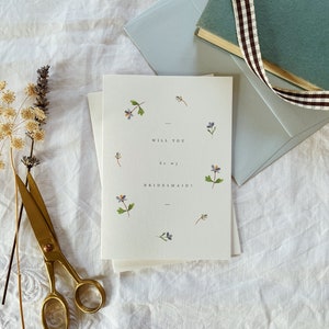 May include: A set of white cards with the text "WILL YOU be my BRIDESMAID!" and floral illustrations. A pale blue envelope, a small book with a brown and white ribbon, and gold scissors are also in the image.