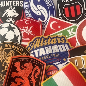Op de afbeelding: Een verzameling kleurrijke geborduurde patches met verschillende ontwerpen en tekst. Patches bevatten logo's van sportteams, vlaggen en grafische illustraties. De patches hebben verschillende vormen, waaronder cirkels, schilden en rechthoeken.