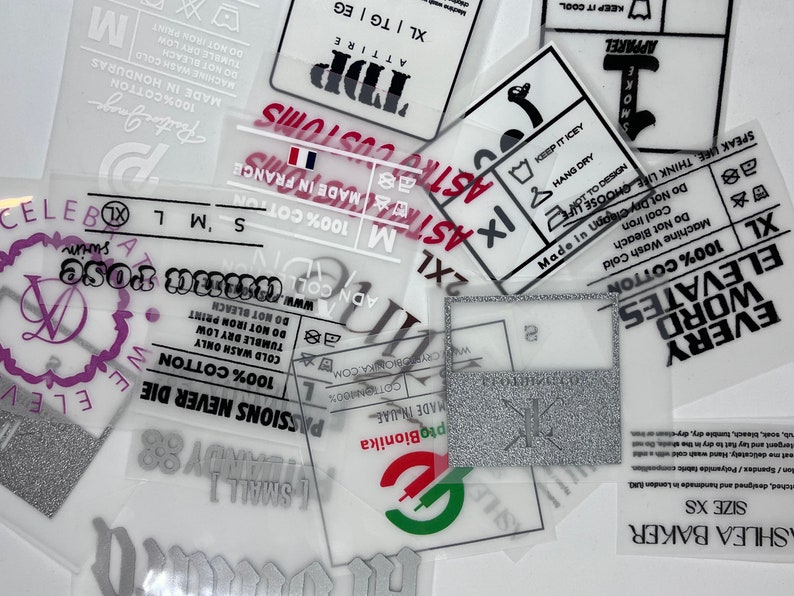 May include: A collection of transparent clothing labels with various text and symbols. The labels are printed with black, white, red, and green ink. The text includes "100% Cotton", "Made in France", "Every Word Elevates", "Astro Customs", "Keep it Icy", "Size XS", "Shlea Baker", and "Passion Never Die".