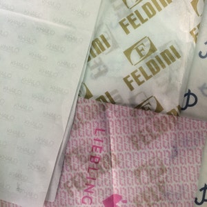 May include: Overhead shot of various tissue papers with printed logos and text. One paper has the name "KHAILO" repeated, another has "FELDINI" in gold, and a third has "LIEBLINGSST" with a heart symbol. The papers are white and pink.