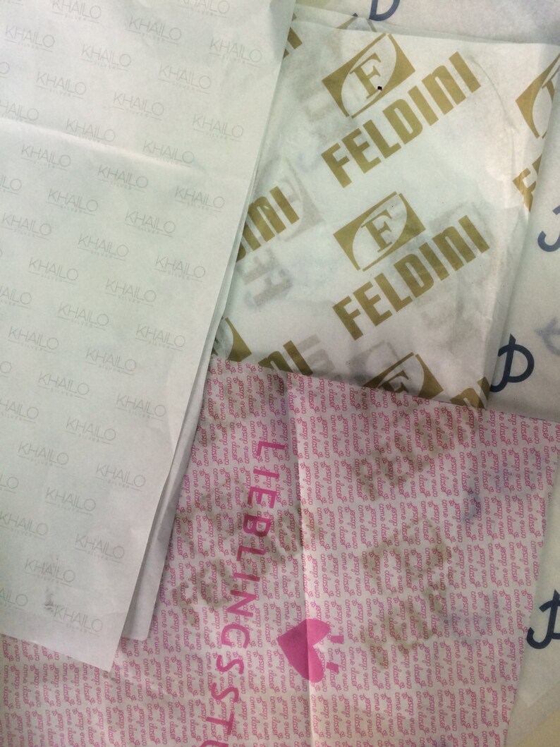 May include: Overhead shot of various tissue papers with printed logos and text. One paper has the name "KHAILO" repeated, another has "FELDINI" in gold, and a third has "LIEBLINGSST" with a heart symbol. The papers are white and pink.