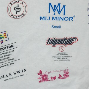 May include: White t-shirt with blue text that reads "PLAY A PLAYER" in a circle, "M M MIJ MINOR" and "Small". The shirt also has a blue and red logo that reads "taigastyle" with a size "S" in a circle. The shirt has a pink floral design and text that reads "Eylul design". The shirt also has text that reads "COTTON", "100% COTTON", "MADE IN CAMBODIA", "MACHINE WASH COLD INSIDE OUT", "TUMBLE DRY LOW", "DO NOT IRON DECORATION", "SHAN SWIN", "HAND WASH COLD ONLY".