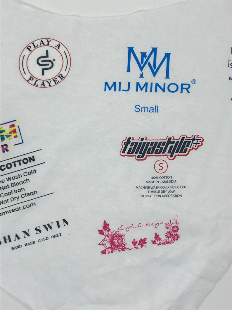 May include: White t-shirt with blue text that reads "PLAY A PLAYER" in a circle, "M M MIJ MINOR" and "Small". The shirt also has a blue and red logo that reads "taigastyle" with a size "S" in a circle. The shirt has a pink floral design and text that reads "Eylul design". The shirt also has text that reads "COTTON", "100% COTTON", "MADE IN CAMBODIA", "MACHINE WASH COLD INSIDE OUT", "TUMBLE DRY LOW", "DO NOT IRON DECORATION", "SHAN SWIN", "HAND WASH COLD ONLY".
