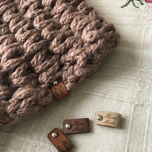 Puede incluir: Primer plano de un artículo de punto marrón con un patrón trenzado texturizado. Varias etiquetas de cuero pequeñas con las palabras "Hand Made" y "Hand with" están esparcidas alrededor del artículo de punto. Las etiquetas están sujetas con remaches plateados.