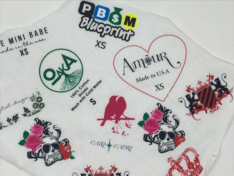 May include: A white fabric with various colorful patches and logos, including "PBSM Blueprint", "OKA", "Amour Made in U.S.A", "CARLO CAPRI", and a skull with roses. The text "E MINI BABE made in the usa XS" is also visible.