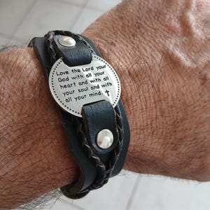 May include: A black leather bracelet with a braided design and a silver plate. The plate has the engraved text: "Love the Lord your God with all your heart and with all your soul and with all your mind."