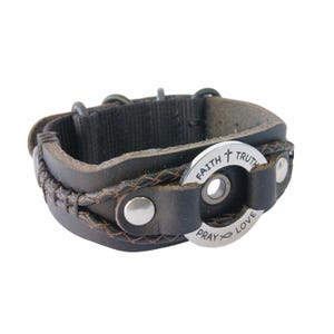 May include: Dark brown leather bracelet with a braided edge and a black fabric backing. A silver-colored circular charm is attached, engraved with the words "FAITH TRUTH PRAY LOVE" and a cross and fish symbol. The bracelet has metal rivets and a buckle closure.