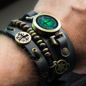 May include: A multi-layered leather bracelet with a variety of embellishments. The bracelet features a central circular charm with the text "I CAN DO ALL THINGS THROUGH CHRIST" and a cross. Additional elements include wooden beads, a tree of life charm, and a star of David charm.