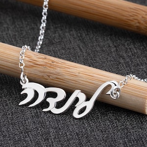 May include: A silver necklace with a cursive name pendant. The necklace is displayed on a wooden dowel against a dark gray background. The chain is delicate and the pendant is in a stylized font.