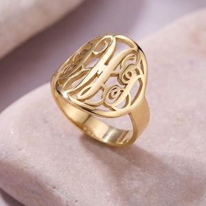 May include: A gold-colored monogram ring with an oval face. The ring features a cut-out design of intertwined letters. The band is smooth and the ring is set against a soft, neutral background.